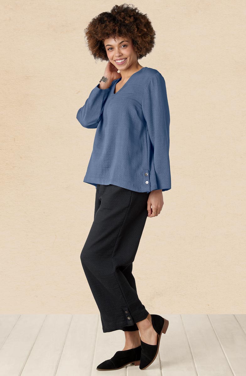 Woman wearing a blue top and black pants against a beige wall.