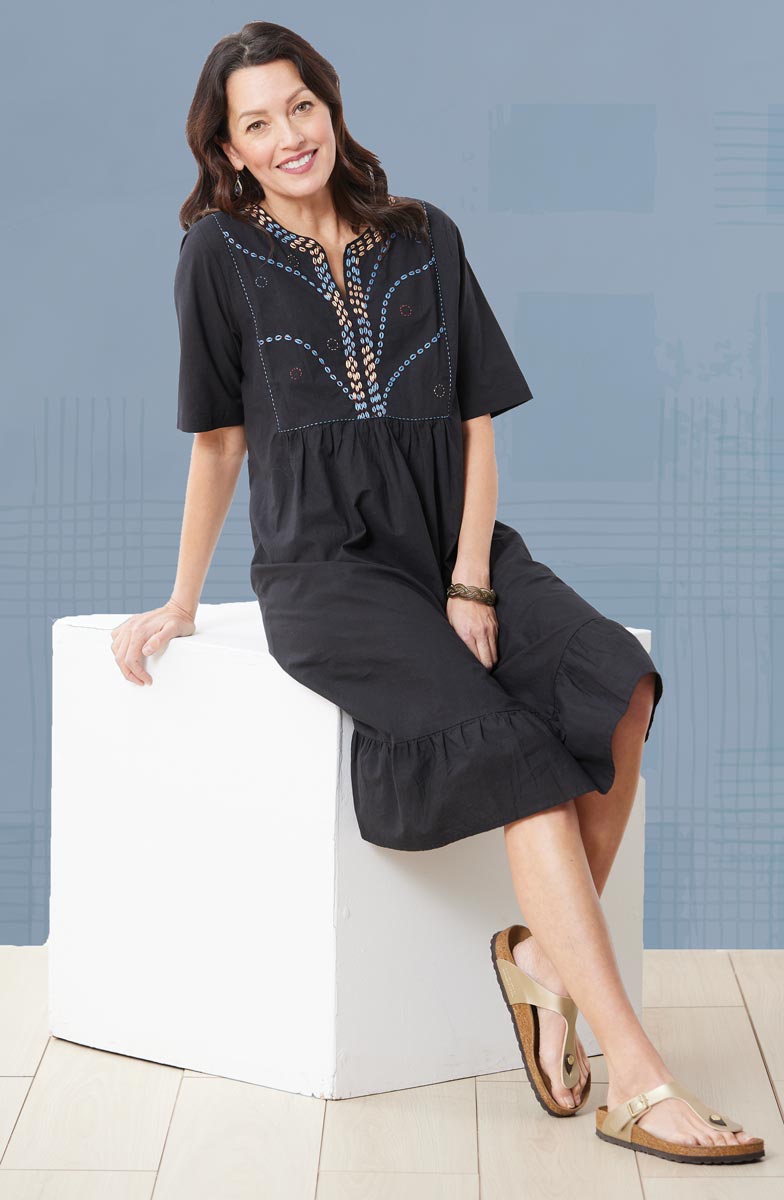 Woman wearing a black dress with embroidery sitting on a white stool against a blue wall.