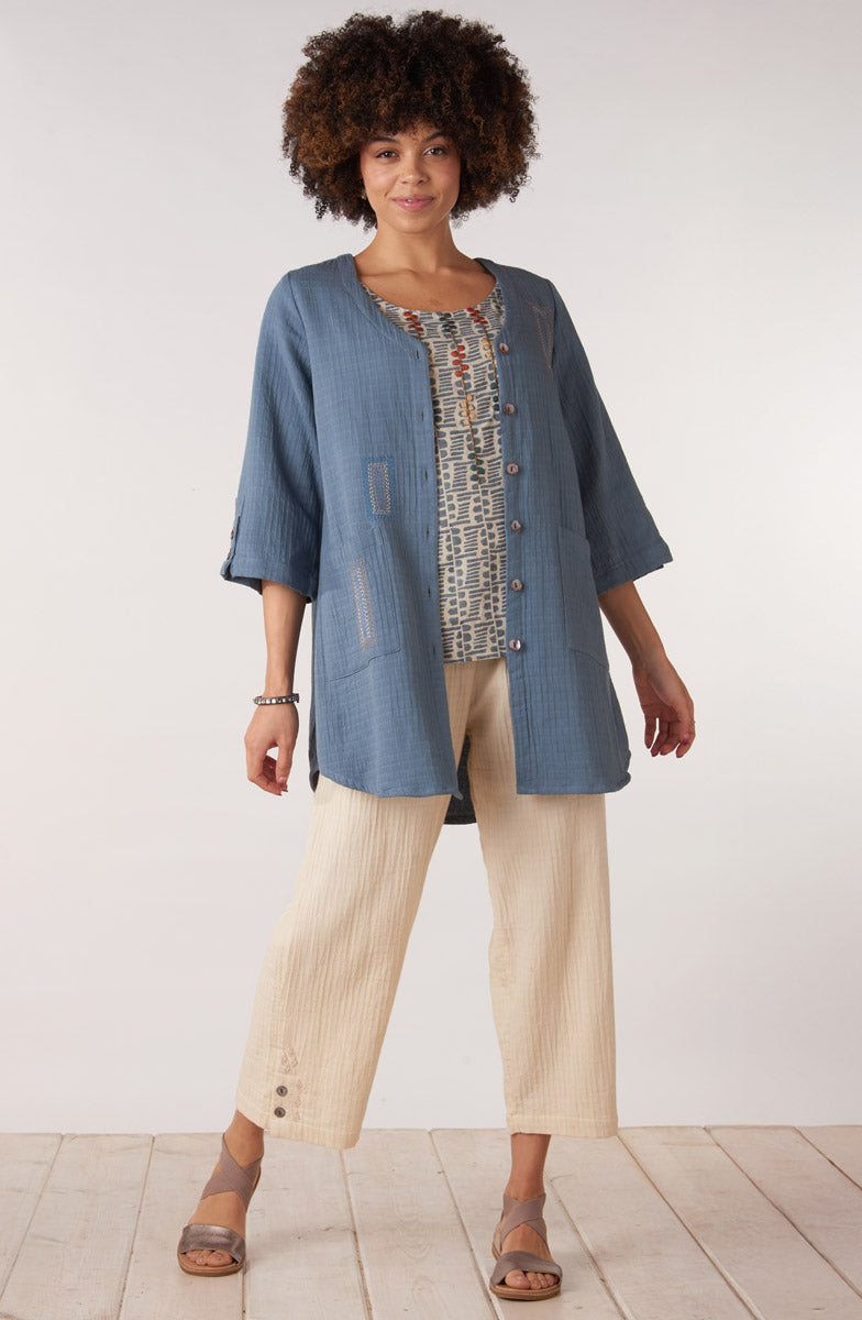 Woman wearing a blue denim jacket over a patterned top with beige pants on a white background