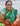 A woman wearing a green and gold saree with orange and red patterns, holding a needle in her hand against a white corrugated metal background.